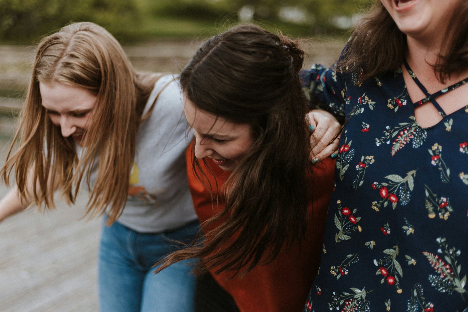 Three women, all friends, standing outside with arms over one another's shoulders. They are laughing and smiling.